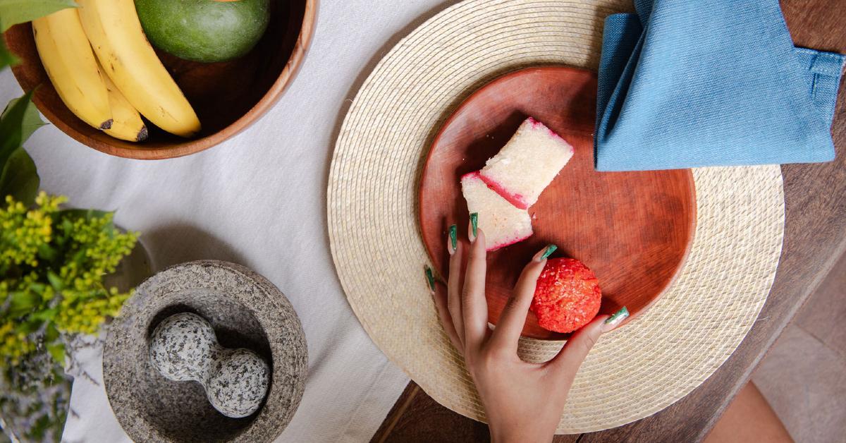 Photo of a manicured hand grabbing food from a plate on a table also showcasing a molcajete, a bowl of fruit, and a cloth napkin. 