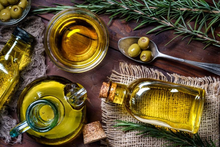 Bottles of olive oil are pictured beside a bowl of olive oil and a spoon with olives on it atop a wooden table.